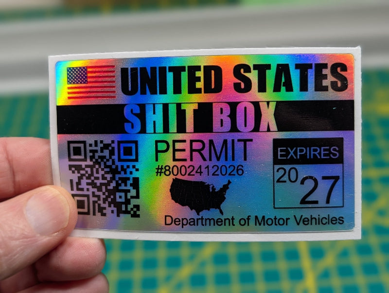 Holographic sticker with text 'UNITED STATES SHIT BOX PERMIT' held by a hand against a blurred background.
