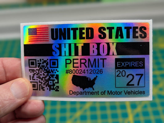 Holographic sticker with text 'UNITED STATES SHIT BOX PERMIT' held by a hand against a blurred background.