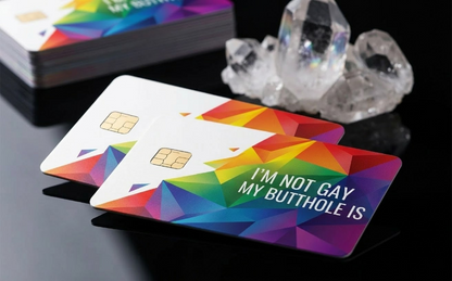 A professional close-up photograph of two modern custom debit cards featuring a cascading rainbow gradient of geometric polygons. White right-aligned text explicitly states "I'M NOT GAY MY BUTTHOLE IS". The cards, which include a visible chip, rest fanned out on a dark reflective surface next to natural quartz crystals.